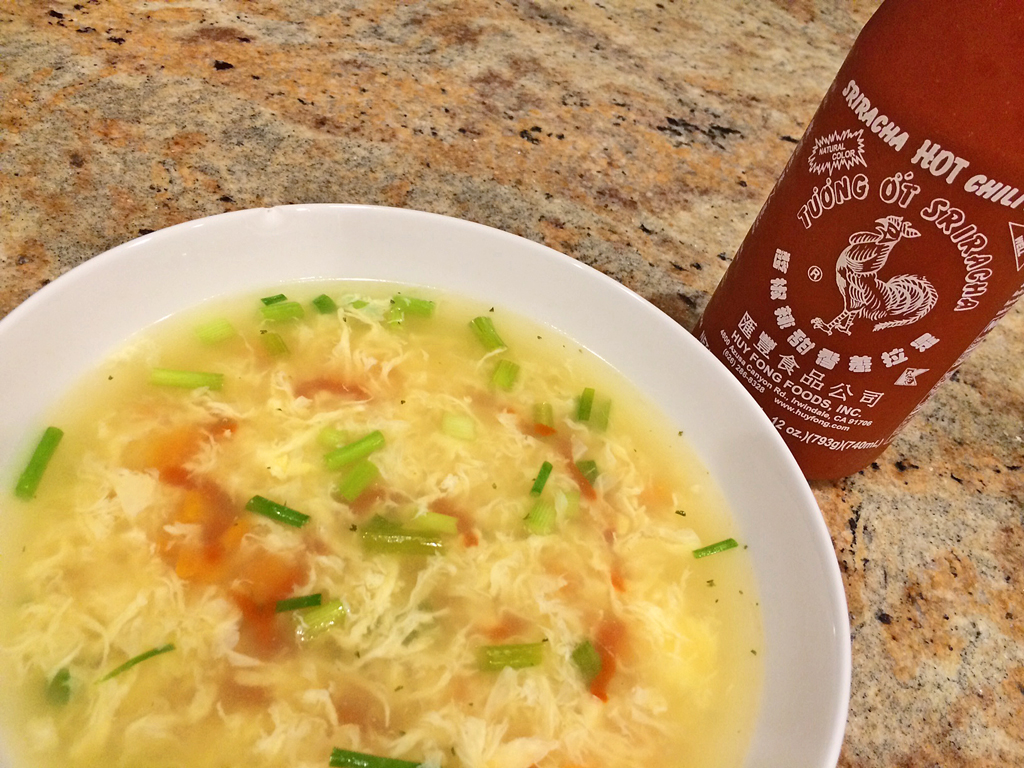 Egg Drop Sriracha Soup in white bowl with green onions and sriracha bottle.